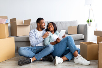 New home ownership. Loving black family using digital tablet and embracing while sitting on floor in new house among unpacked moving boxes, copy space. Real estate concept
