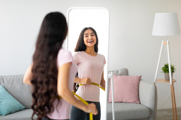 Attractive young Indian woman measuring her waist with tape near mirror indoors, copy space. Young...