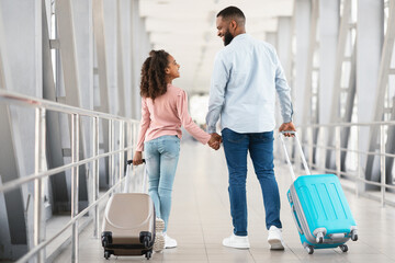 Ready For Travel. Rear back view of happy black father and daughter walking in modern airport corridor, carrying suitcases at terminal. Smiling parent holding hands with daughter looking at each other
