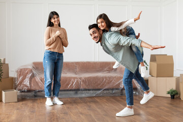 Having Fun On Moving Day. Full body length of cheerful father carrying his daughter on back spreading arms as wings pretending flying, imitating air plane with hands, mum standing in empty living room © Prostock-studio