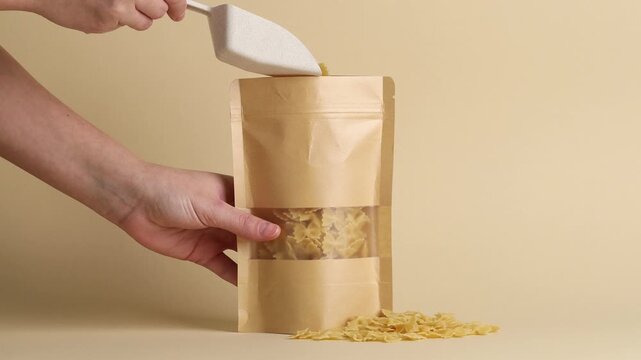 Woman putting raw pasta into stand-up pouch on beige background, closeup