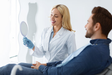 Obraz premium Pretty young blonde woman dentist holding mirror for male patient sitting in dental chair, stomatologist showing results of treatment, cheerful barded man looking at mirror and smiling, copy space