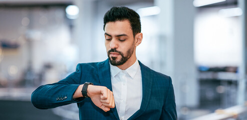 A man in a suit is looking at his watch while standing in an office. The background shows an open workspace with desks and computers. He appears focused and attentive to time. © Prostock-studio