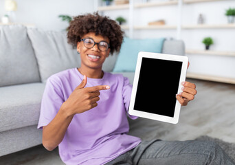 Cheerful black teenager pointing at tablet pc with blank screen at home, mockup for mobile app or website design. Online education, remote communication during coronavirus isolation