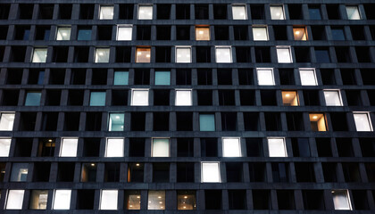 Modern dark concrete building facade at night with a symmetrical window grid, where only some office lights are illuminated, suggesting corporate overtime.