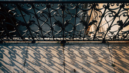Top-down view of ornate wrought iron railing casting long, decorative shadows onto a textured stone balcony floor in strong sunlight.
