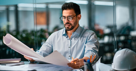 A man sits at a desk in an office, closely examining blueprints and plans. He is focused on the details. A hard hat and tools are visible on the desk, indicating a construction project.