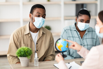 Black Tourists Couple And Travel Agent Lady Choosing Destination For Vacation Pointing At Globe Sitting In Tour Agency Office Indoors, Wearing Face Masks. Selective Focus