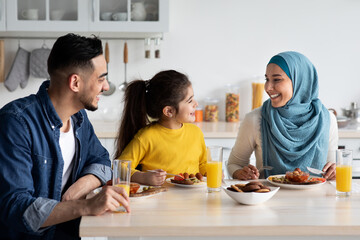 Fototapeta premium Domestic Breakfast. Happy Middle Eastern Family Of Three Eating Together In Kitchen, Cheerful Young Arab Parents And Little Daughter Sitting At Table, Enjoying Tasty Food, Chatting And Laughing