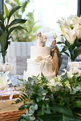 Elegant wedding dessert table with white tiered cake and lily floral arrangements, sophisticated reception decor with sweets display and religious icon, bright event venue interior setting