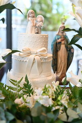 Three-tiered white wedding cake with bow and figurines, elegant reception dessert table with Virgin Mary statue and white lilies, sophisticated marriage celebration cake with floral decor