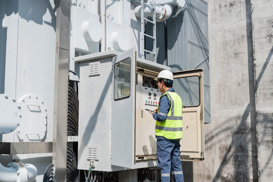 Electrical engineer in safety vest using digital tablet to inspect control panel of high voltage transformer at substation, monitoring energy system performance and industrial infrastructure.