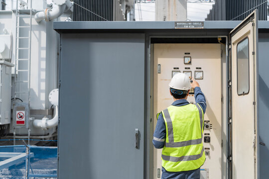 Electrical engineer in safety vest pointing and inspecting control panel gauges inside Marshalling Cubicle at power substation, monitoring industrial energy system for grid maintenance.