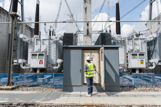 Male electrical engineer inspecting control cabinet of large power transformers at high voltage substation to monitor energy distribution grid and industrial utility infrastructure.