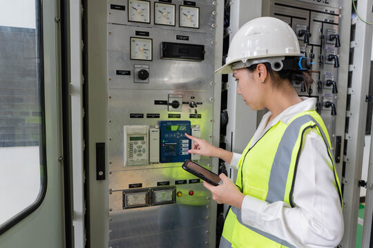 Female electrical engineer tests digital protection relay on control panel, verifying system parameters and operational status to ensure power reliability and safe operation.