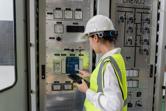 Female electrical engineer operating protection relay and control system on indoor panel while holding digital tablet for maintenance audit at high voltage substation facility.