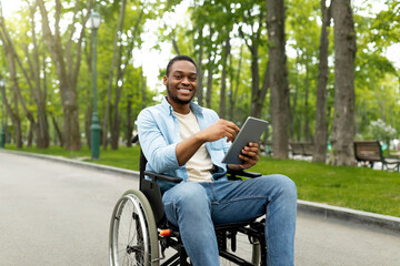Fototapeta premium Portrait of cheeful impaired young man in wheelchair using laptop for remote communication at city park. Happy black guy with disability having online meeting, chatting with friend on web outdoors