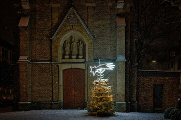 Atmospheric winter night in Cologne Ehrenfeld. A glowing Christmas tree with a shooting star topper stands in the snow before the historic Marktkapelle chapel