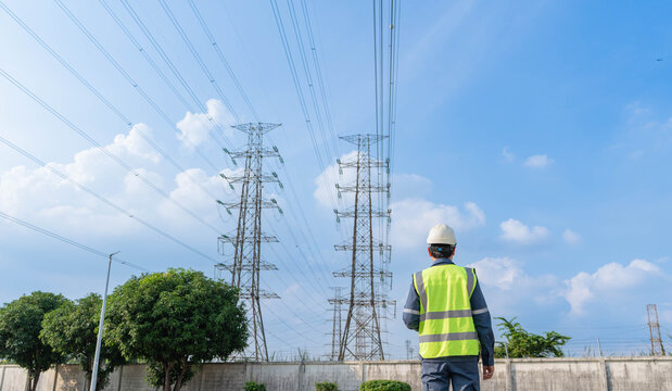 Back view of electrical engineer looking at high voltage power line pylon under blue sky. Maintenance worker planning grid infrastructure development and energy distribution.