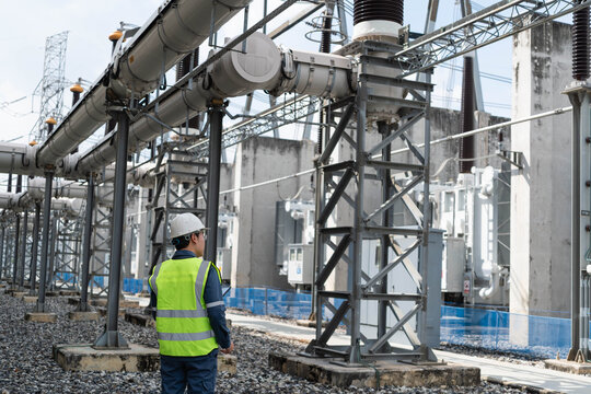 Professional electrical engineer inspecting outdoor high voltage substation infrastructure and power transformers. Technician in safety vest monitoring energy grid equipment.