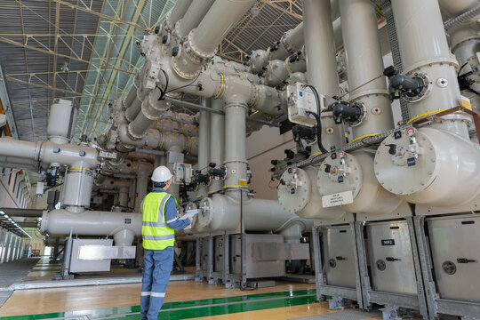 Wide angle view of electrical engineer standing among massive gas insulated switchgear pipes in industrial substation. Professional maintenance of high voltage energy infrastructure.