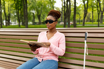 Millennial black visually impaired woman sitting on bench at city park, reading Braille book outside. Young blind lady in dark glasses studying outdoors. Education and disability concept