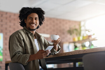 Cheerful millennial african american curly man with smartphone sit at table, drink cup of coffee and look at camera in cafe interior. Social networks and chat, modern device and blog at spare time