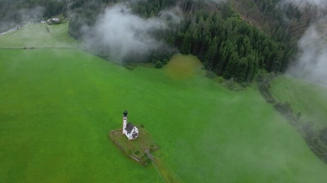 aerial solitary steeple rising from green expanse heavy fog swirling around meadow cinematic drone sweep slow reveal pastoral atmosphere ideal for aerial broll travel documentaries