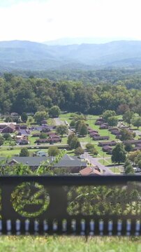 Verti al screen aerial suburban panorama Johnson City overlook showcasing grid houses, blue mountain ridges, ornamental park railing with emblem, bright midday sun, smooth drone glide, high