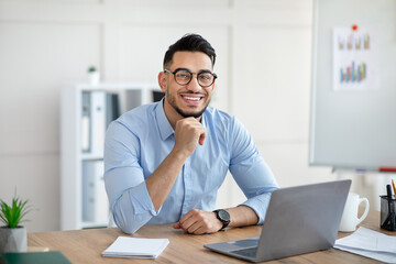 Obraz premium Portrait of happy Arab businessman using laptop computer, making video call, chatting online with colleague at office. Young male entrepreneur having virtual meeting or business webinar