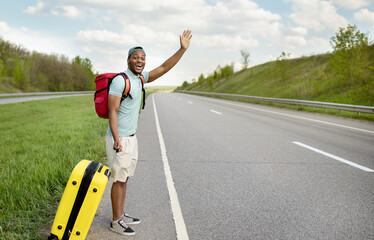 Happy black guy with backpack and suitcase flagging down car, needing ride, hitchhiking on...