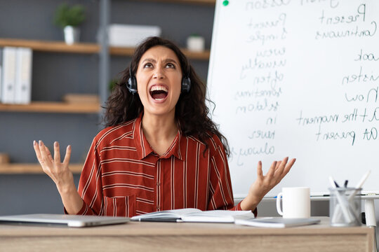Fatigued From Work. Tired young teacher in headset screaming out loud, gesturing and looking up, having video lesson with students, tutoring English language online, sitting at desk and yelling