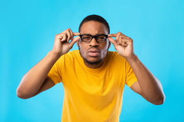Fototapeta premium African American Guy With Poor Eyesight Wearing Eyeglasses And Squinting Eyes Looking At Camera Through Glasses Standing Over Blue Studio Background. Healthcare, Vision Problems