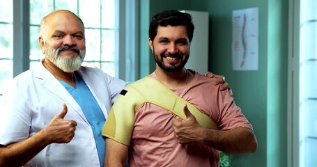 Indian physiotherapist with happy patient with shoulder support smiling and showing thumbs up after therapy as experienced male therapist stands with young man inside modern Indian clinic - Powered by Adobe