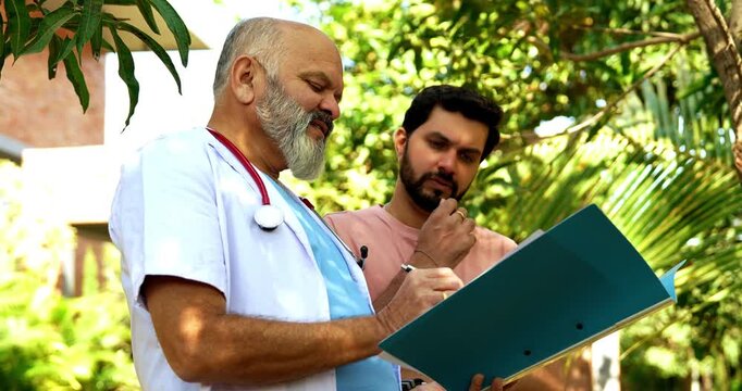 Indian doctor patient discuss case outdoors in natural greenery as senior male physician reviews medical report on laptop or looking at case file with patient during open air healthcare consultation