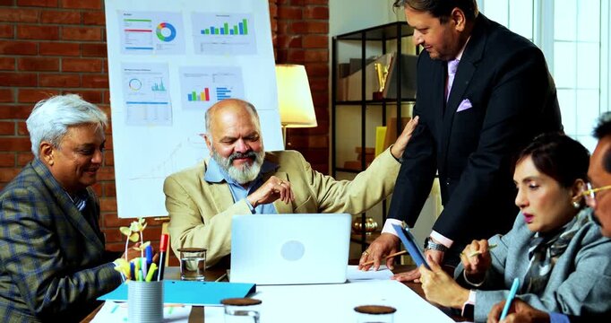 Indian senior business people working in office together as group of senior Indian Asian businessmen collaborate using laptop, papers at desk while analyzing tasks in cozy modern corporate workplace