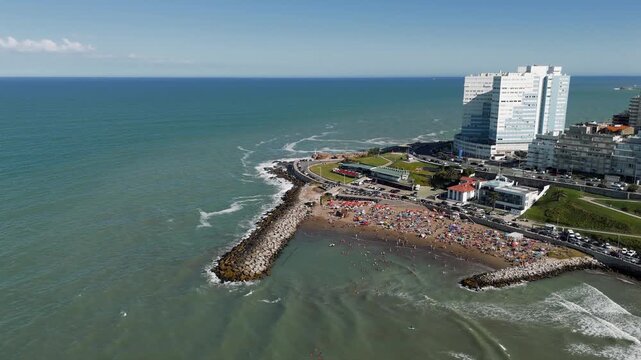 Aerial view of Cabo Corrientes beach in Mar del Plata, Argentina, with people enjoying a sunny summer day. The camera performs an approach, push in, tilt down, and rotate movement