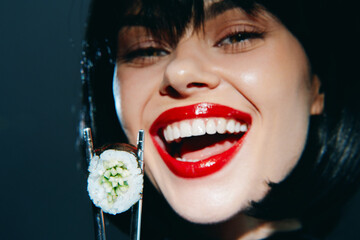 Young woman with red lips joyfully holding sushi with chopsticks, showcasing vibrant colors and modern styling against a dark background.