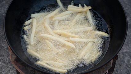 Above view of deep frying french fries in hot boiling oil in a black pan. Crispy potato sticks cooking. Fast food, street food, and unhealthy snack concept.