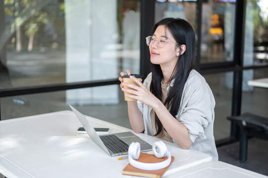 Glasses asian woman college student holding coffee cup over laptop and sitting at table in building.
