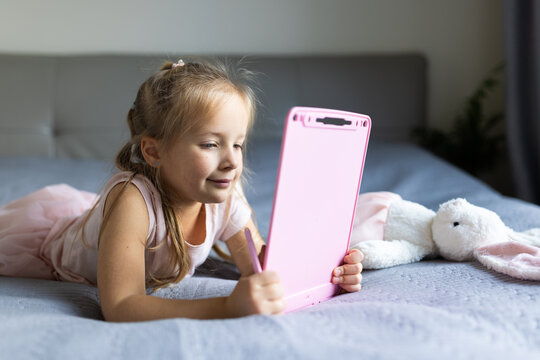 Young girl using a pink drawing tablet while lying on a bed indoors