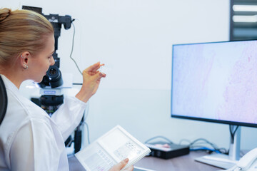 Caucasian female scientist holding glass slide near computer monitor with histology image. Researcher reviewing pathology data in modern medical lab for diagnosis.
