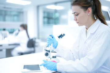 Caucasian female scientist using digital tablet to record data in modern laboratory. Researcher in white coat and blue gloves analyzing results near microscope.