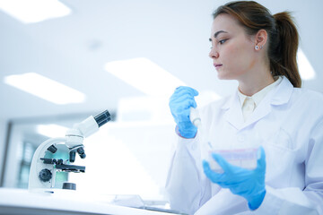Caucasian female scientist using digital tablet to record data in modern laboratory. Researcher in white coat and blue gloves analyzing results near microscope.
