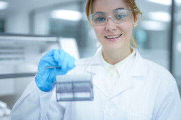 Portrait of a smiling female scientist holding a rack of microscope slides near an automated staining machine. Professional researcher preparing tissue samples for medical pathology diagnosis.
