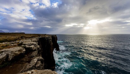 Dramatic coastal landscape with rugged cliffs overlooking a vast ocean under a cloudy sky with sunbeams breaking through.