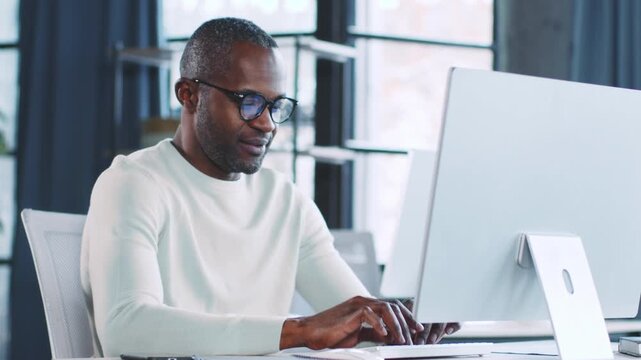 A man works intently at a sleek computer in a stylish office space. Natural light fills the room, enhancing the productive atmosphere. The scene reflects a contemporary business setting.