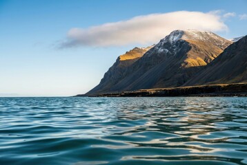 Mountain coastline over calm ocean, snow-capped peaks under blue sky, gentle ripples and golden light, scenic seascape background
