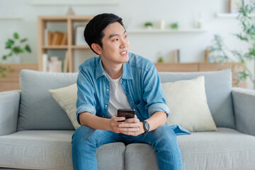 Smiling handsome young Asian man sitting on cozy couch, using funny mobile apps in living room. man at home, doing online shopping, messaging friends, posting on social media