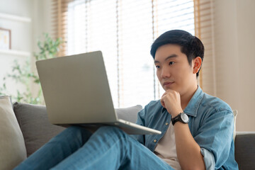 Young Asian man relaxing on comfortable couch, using laptop at home, happy boy chatting with friends in social network, spending weekend, watching movie, shopping online